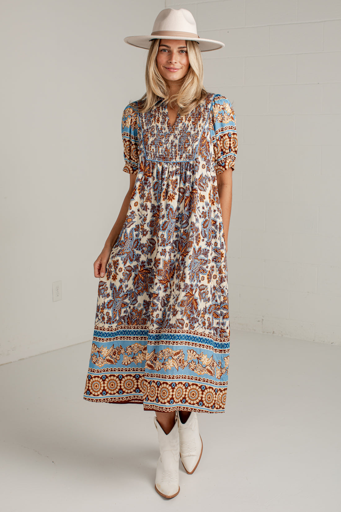 A woman modeling a bohemian-style midi dress with a multicolor paisley and geometric border print, paired with a beige wide-brim hat and white cowboy boots, standing against a white studio wall.