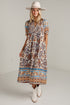 A woman modeling a bohemian-style midi dress with a multicolor paisley and geometric border print, paired with a beige wide-brim hat and white cowboy boots, standing against a white studio wall.