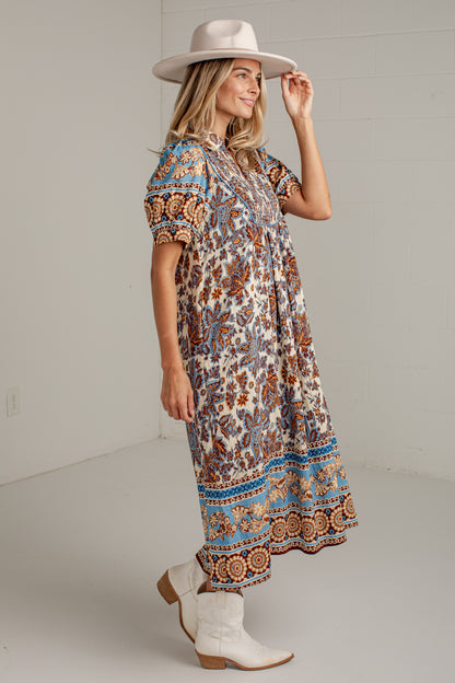 A woman modeling a short-sleeve bohemian maxi dress with a brown, blue, and beige paisley pattern, styled with a beige wide-brim hat and white cowboy boots against a white studio background.