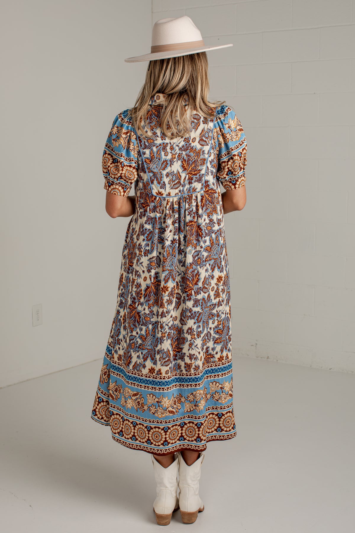 A woman viewed from behind wearing a bohemian-style midi dress with a cream base and blue, brown, and orange floral patterns. She is wearing a wide-brimmed beige hat and white cowboy boots against a white studio background.