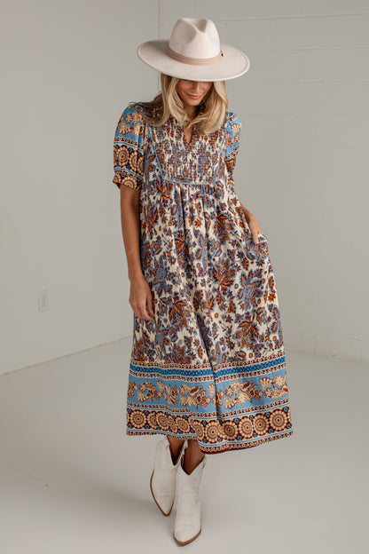 A woman modeling a bohemian-style midi dress with a smocked bodice and a colorful paisley border, paired with a wide-brimmed beige hat and white cowboy boots.