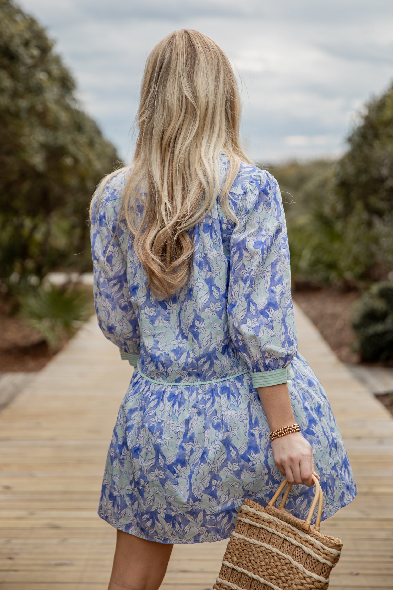 Woman in a blue floral dress holding a straw bag on a wooden path with greenery.