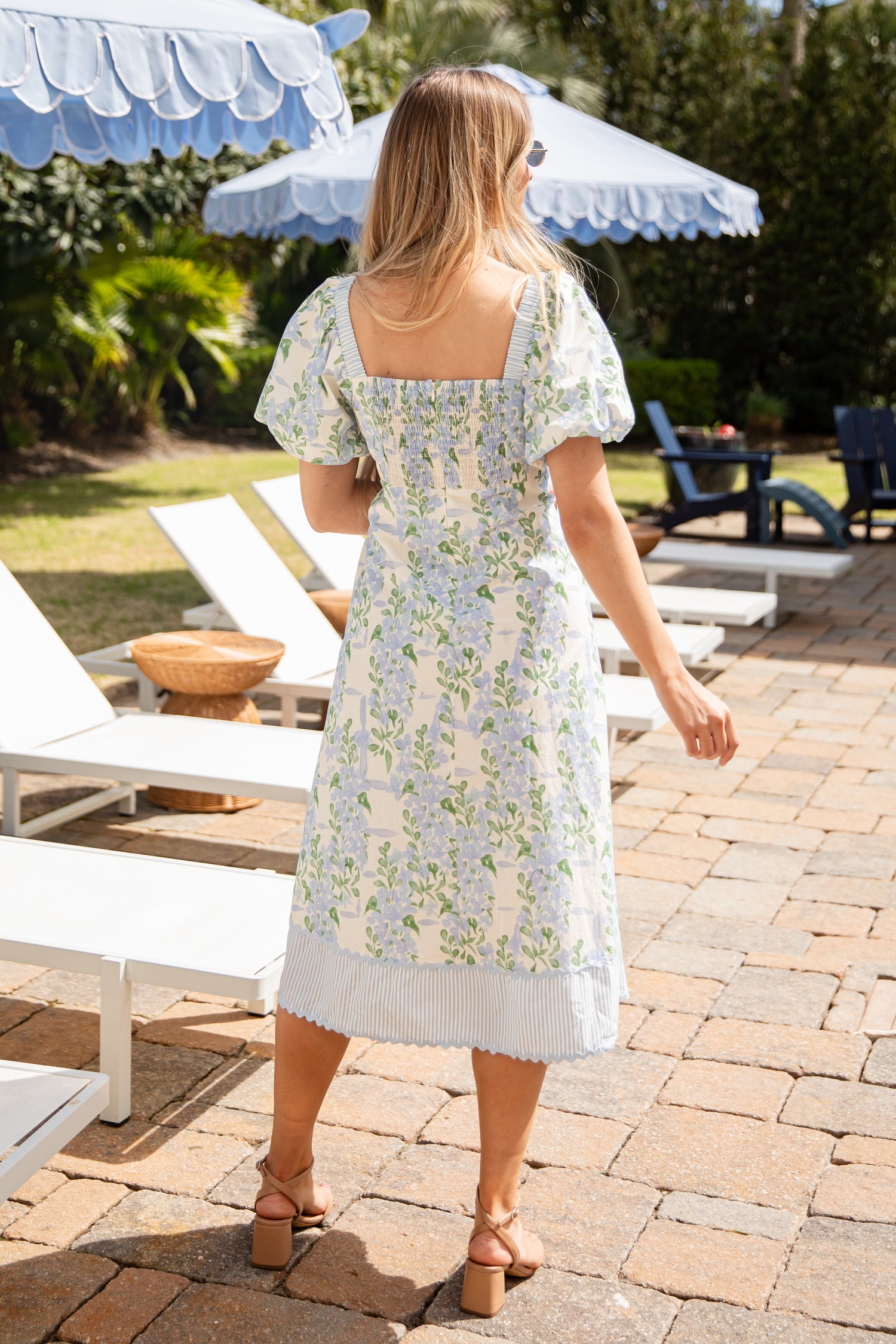Woman in a floral dress standing on a patio with lounge chairs and umbrellas in the background.