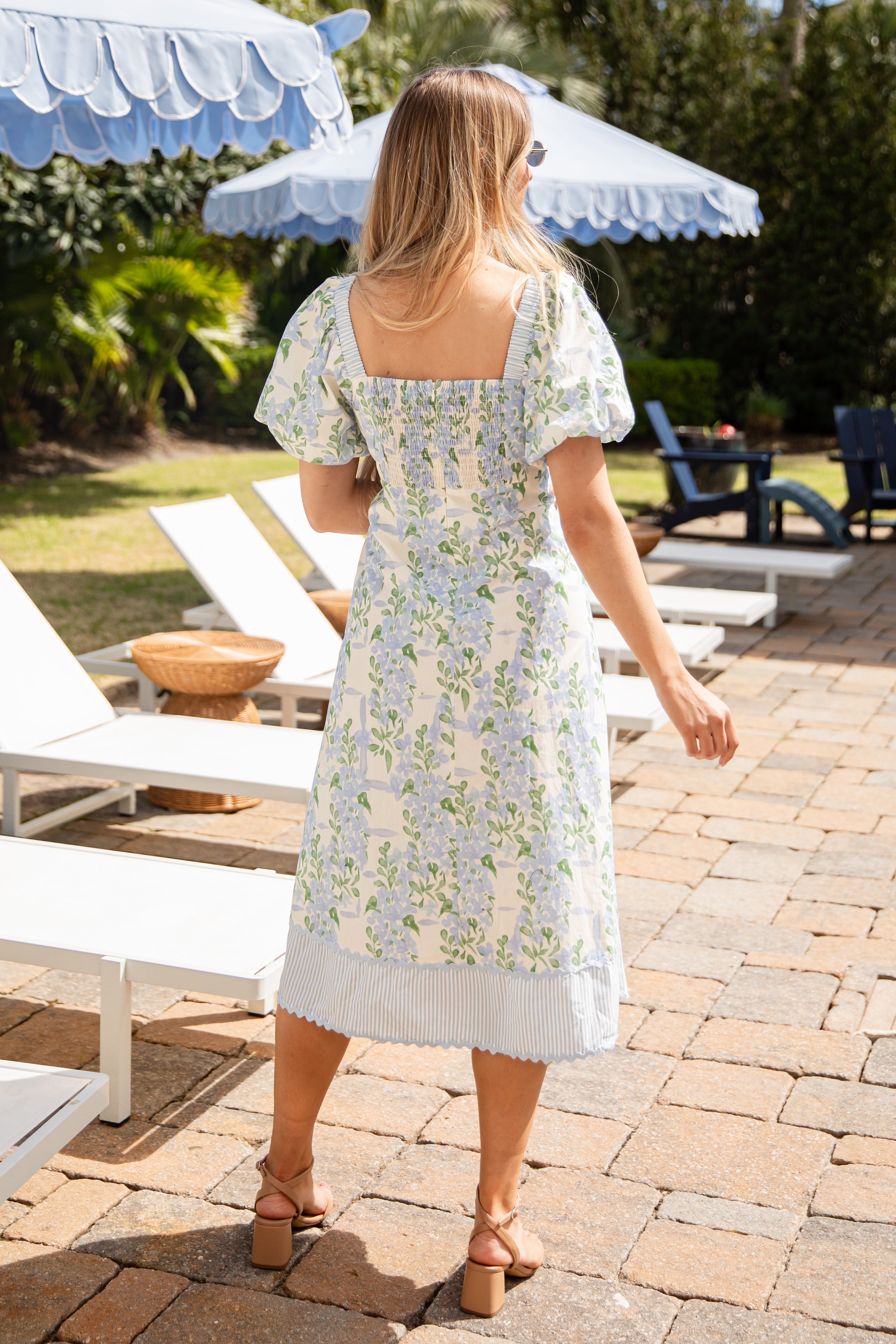 Woman in a floral dress standing on a patio with lounge chairs and umbrellas in the background.