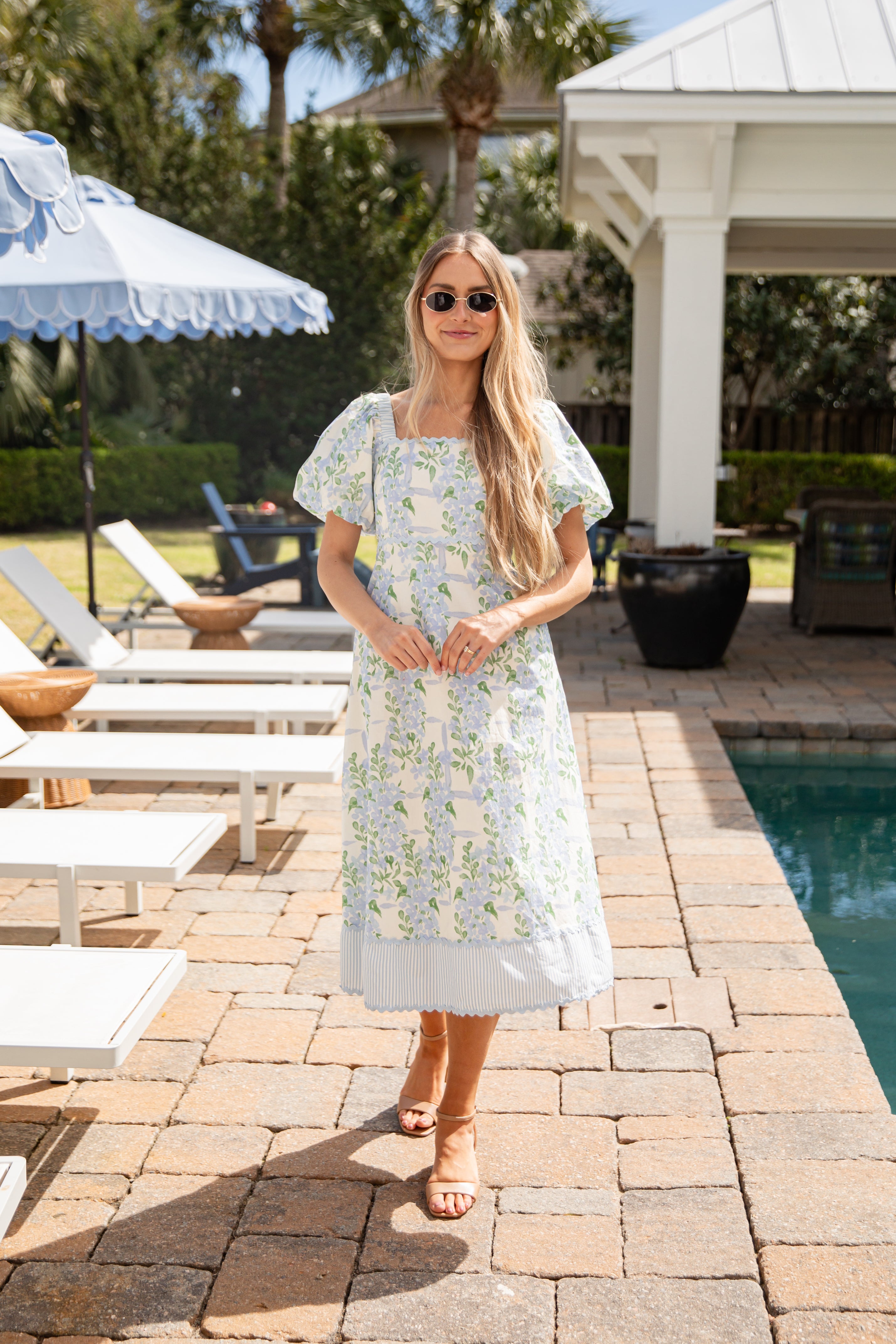 Woman in a floral dress standing by a poolside with lounge chairs and umbrellas.