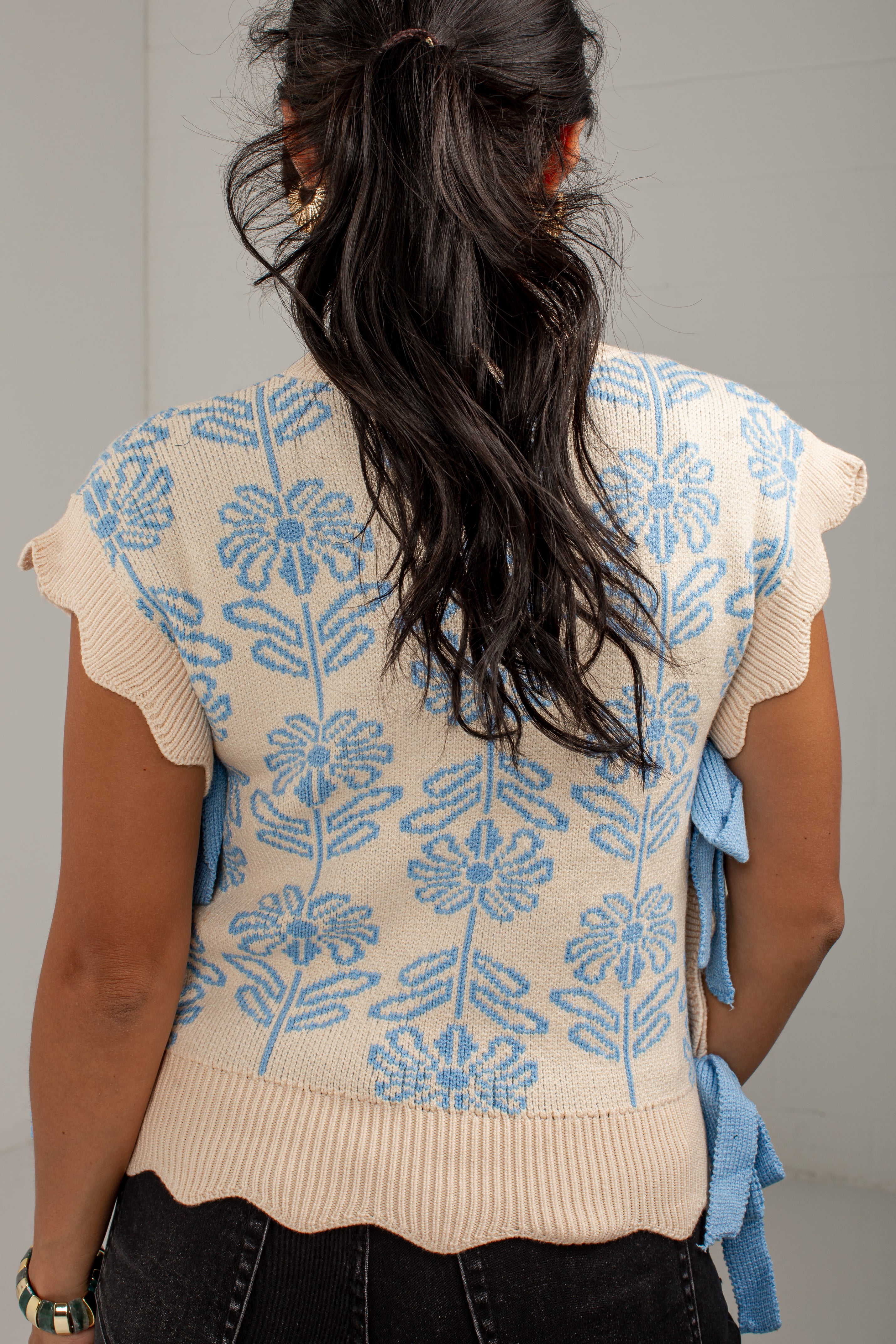 Back view of a woman wearing a cream knit top with a light blue floral pattern, ruffled cap sleeves, and blue side ties, paired with black jeans.