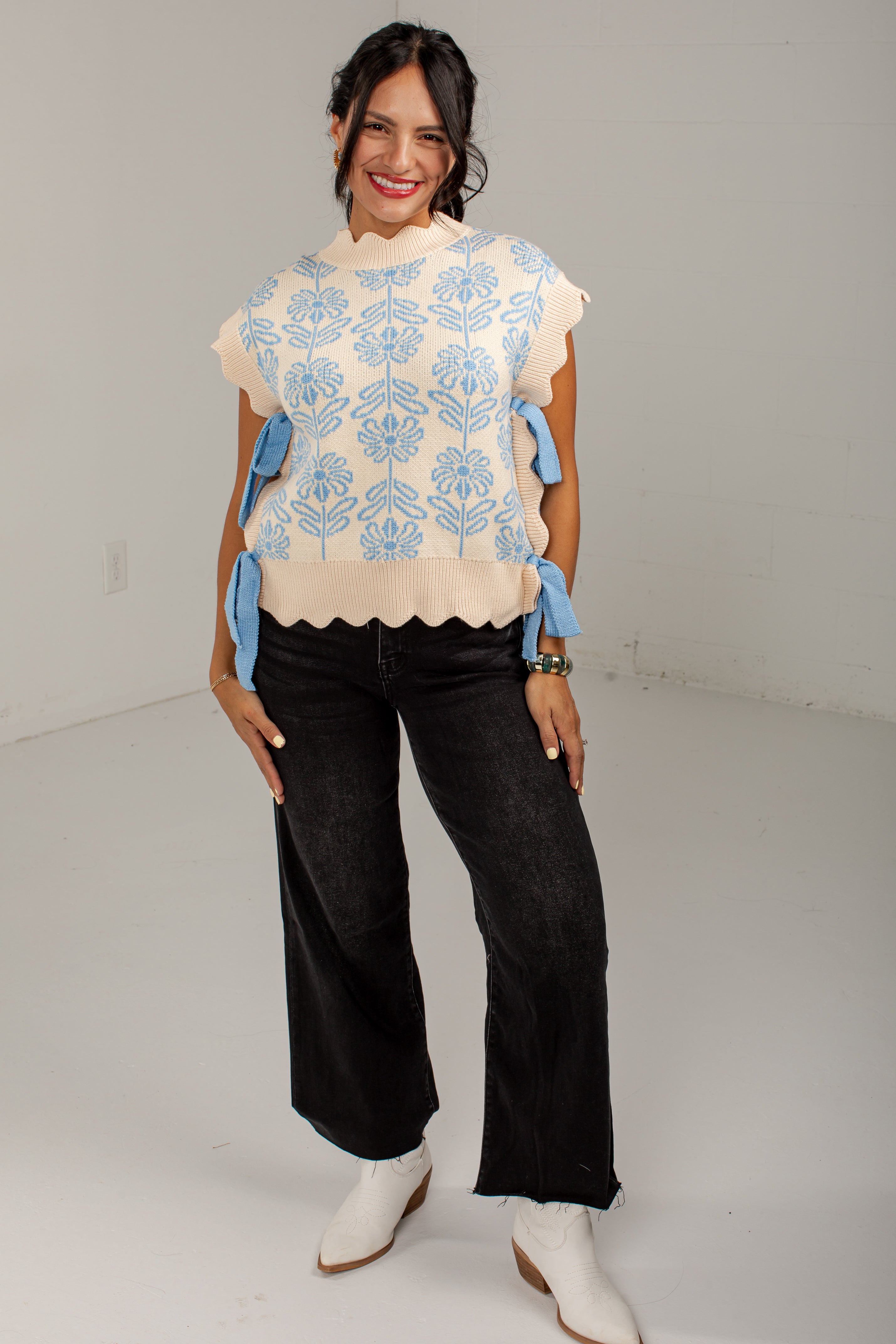 A woman modeling a cream knit top with light blue floral embroidery and blue ribbon tie details on the sleeves, paired with black wide-leg jeans and white cowboy boots, in a white studio setting.