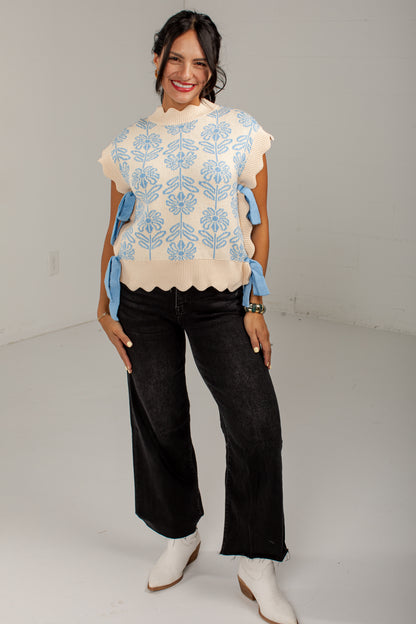 A woman modeling a cream knit top with light blue floral embroidery and blue ribbon tie details on the sleeves, paired with black wide-leg jeans and white cowboy boots, in a white studio setting.