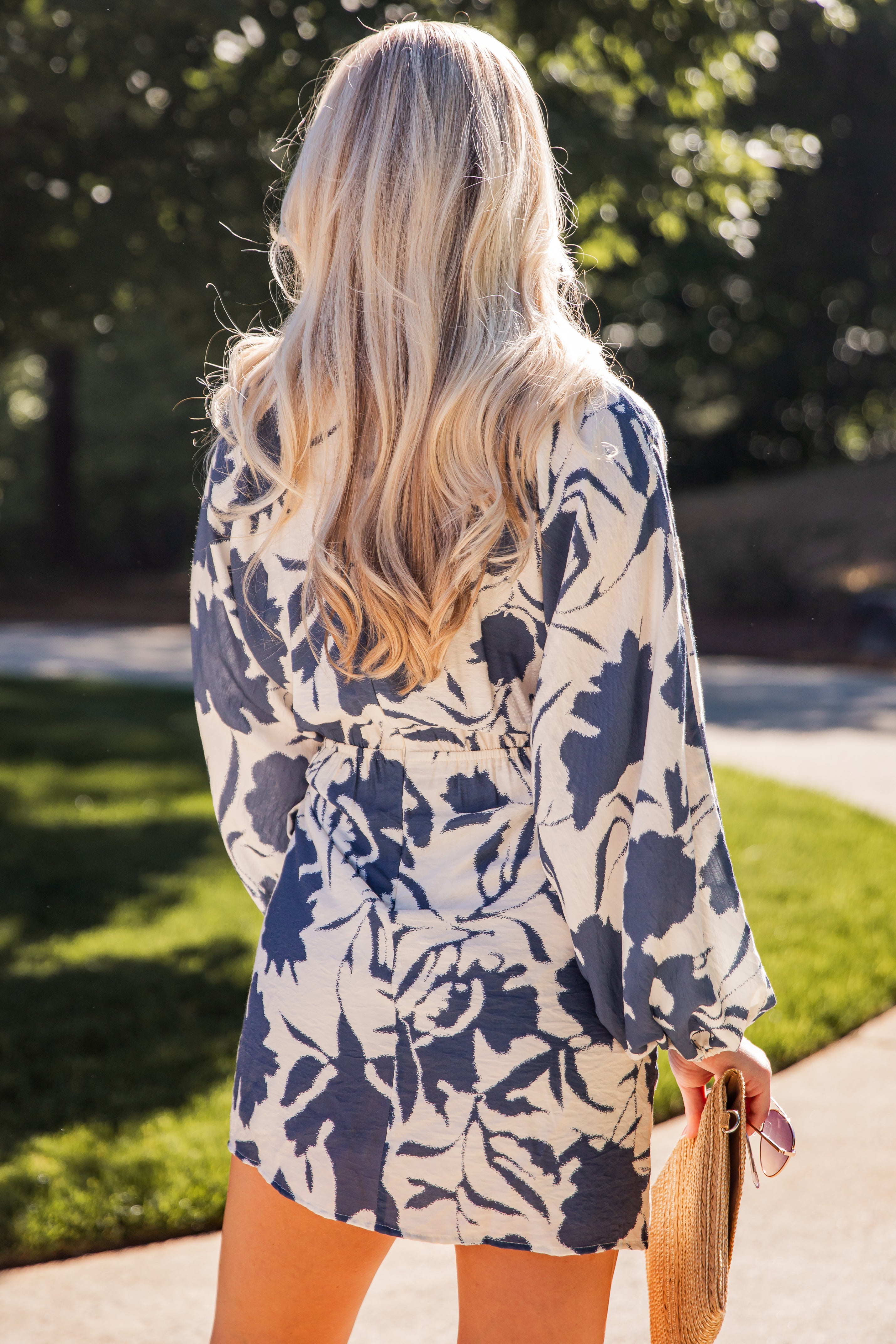 Model wearing a blue floral print wrap mini dress with long sleeves and a waist tie, styled with a woven clutch outdoors.