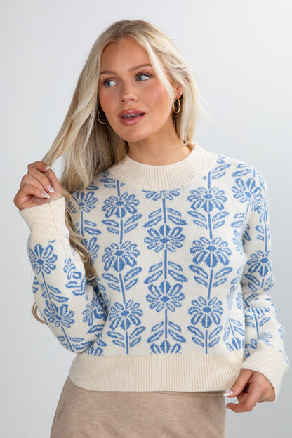 Woman wearing a white sweater with blue floral patterns against a plain background