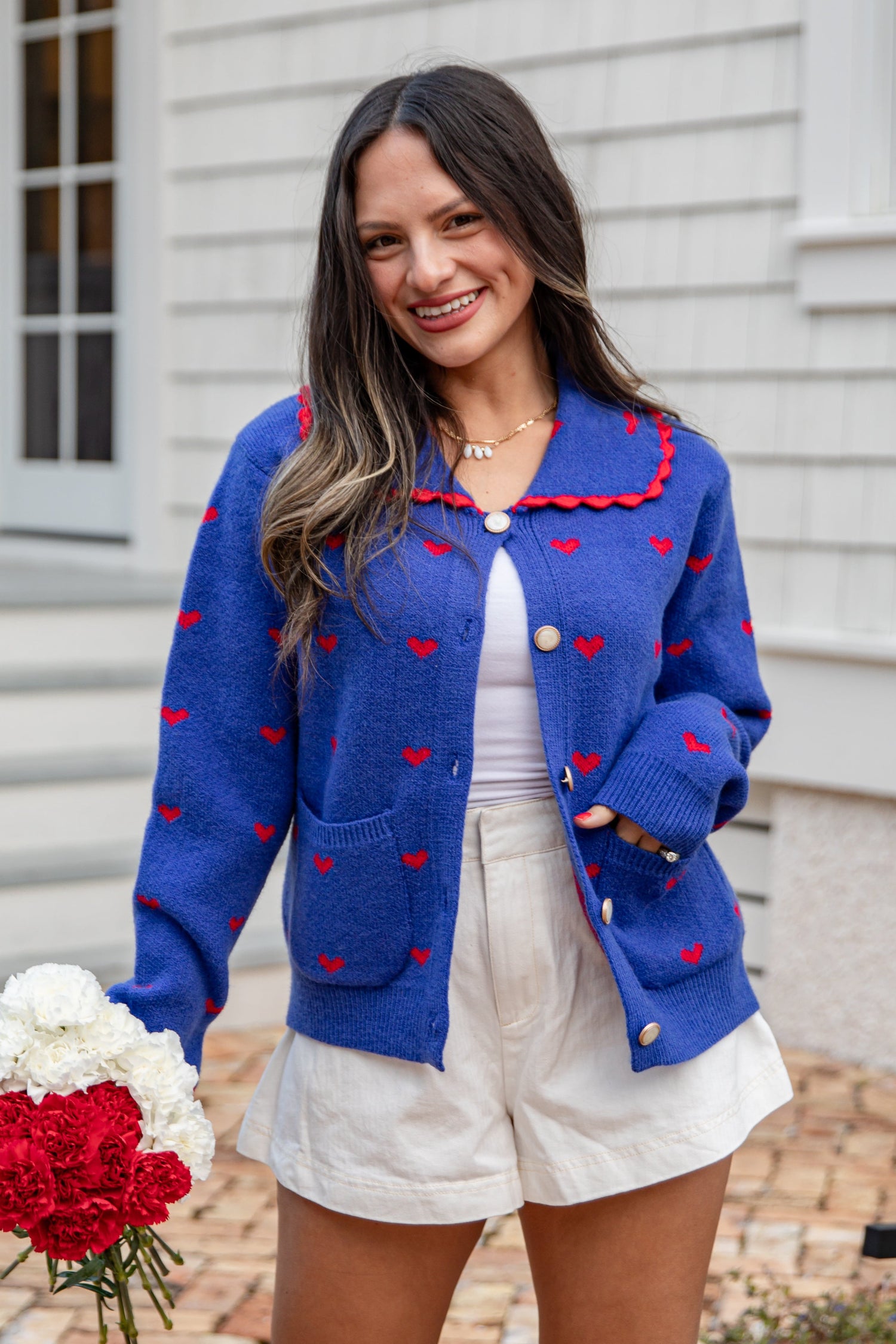 Woman wearing a blue cardigan with red heart patterns, holding flowers, standing in front of a building.