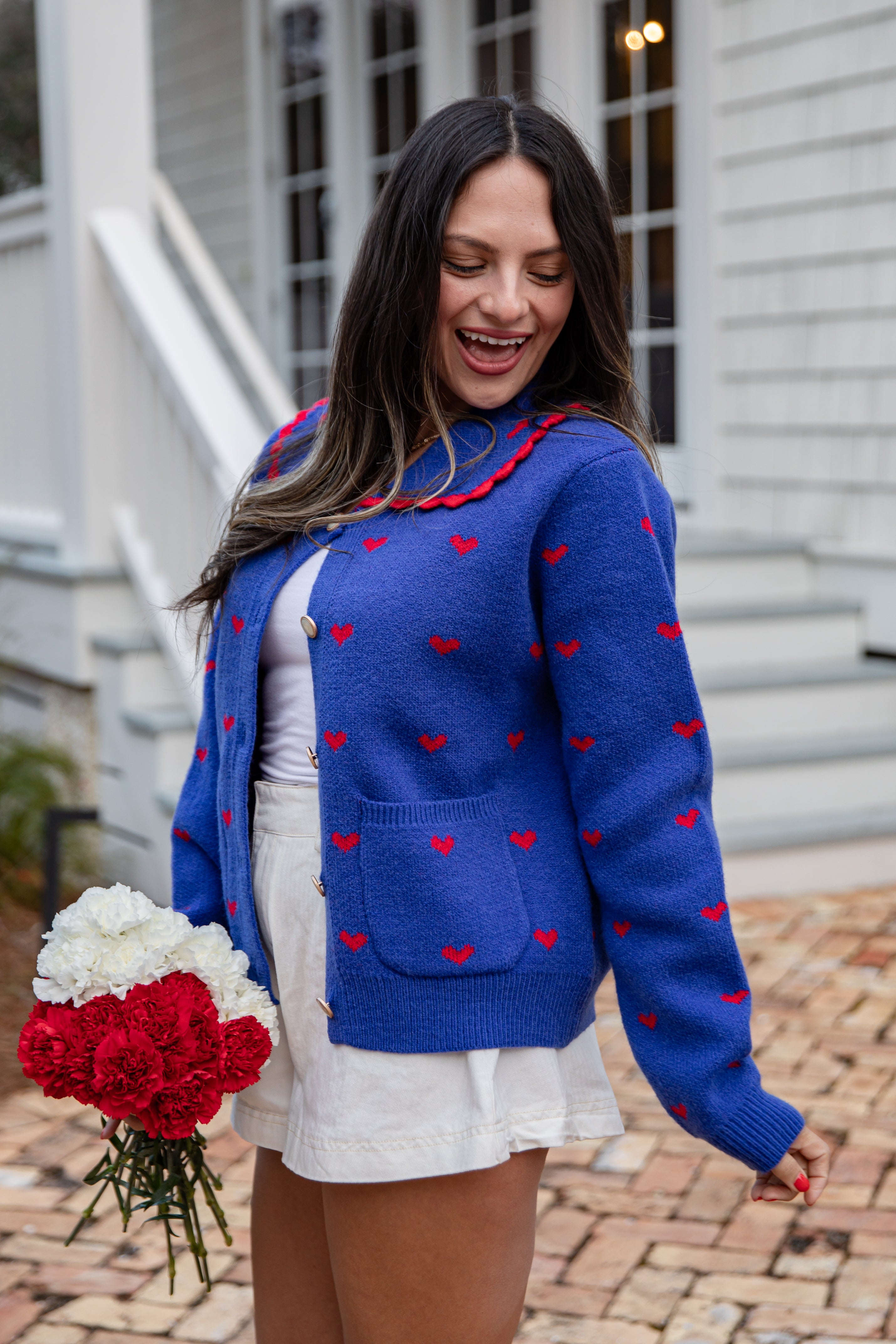 Woman wearing a blue sweater with red heart patterns, holding flowers, standing outdoors.
