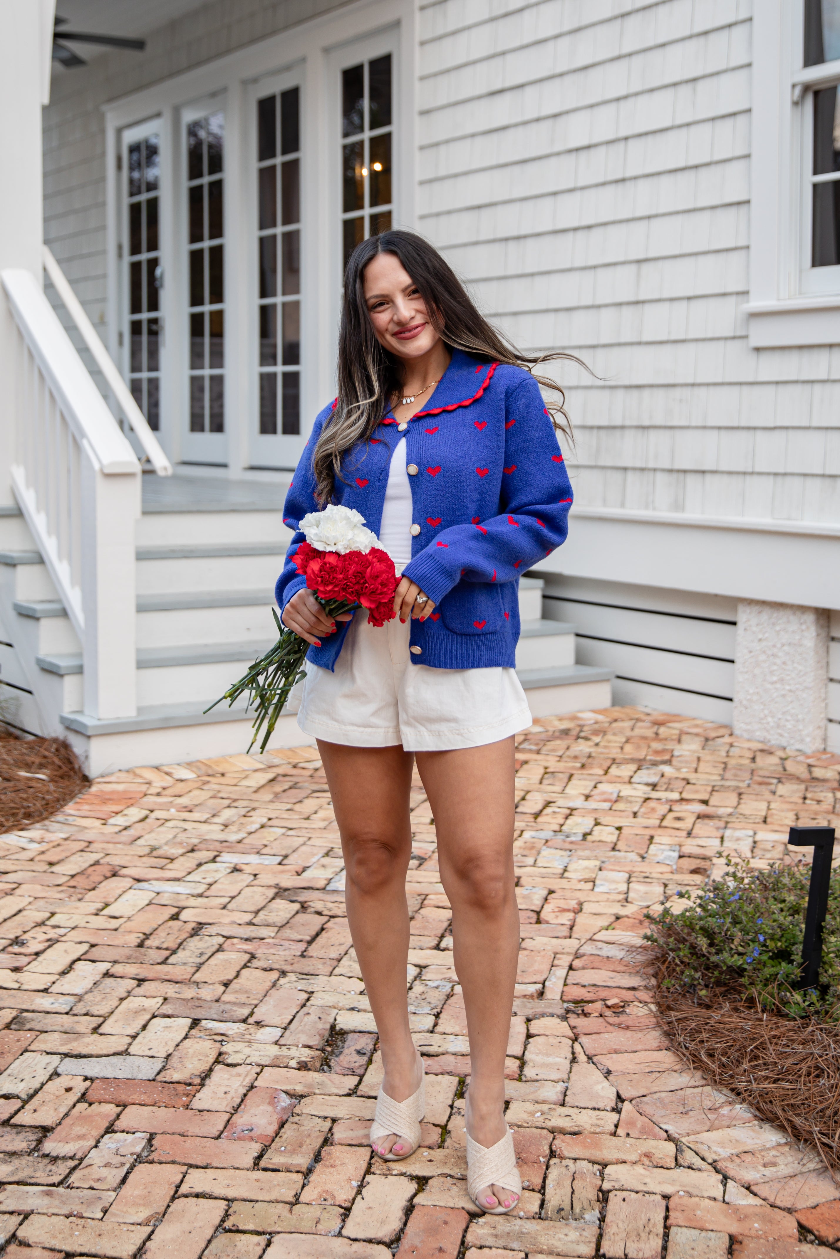 Woman in a blue cardigan with red patterns, white shorts, and sandals holding flowers in front of a house.