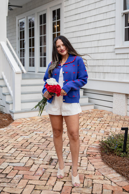 Woman in a blue cardigan with red patterns, white shorts, and sandals holding flowers in front of a house.