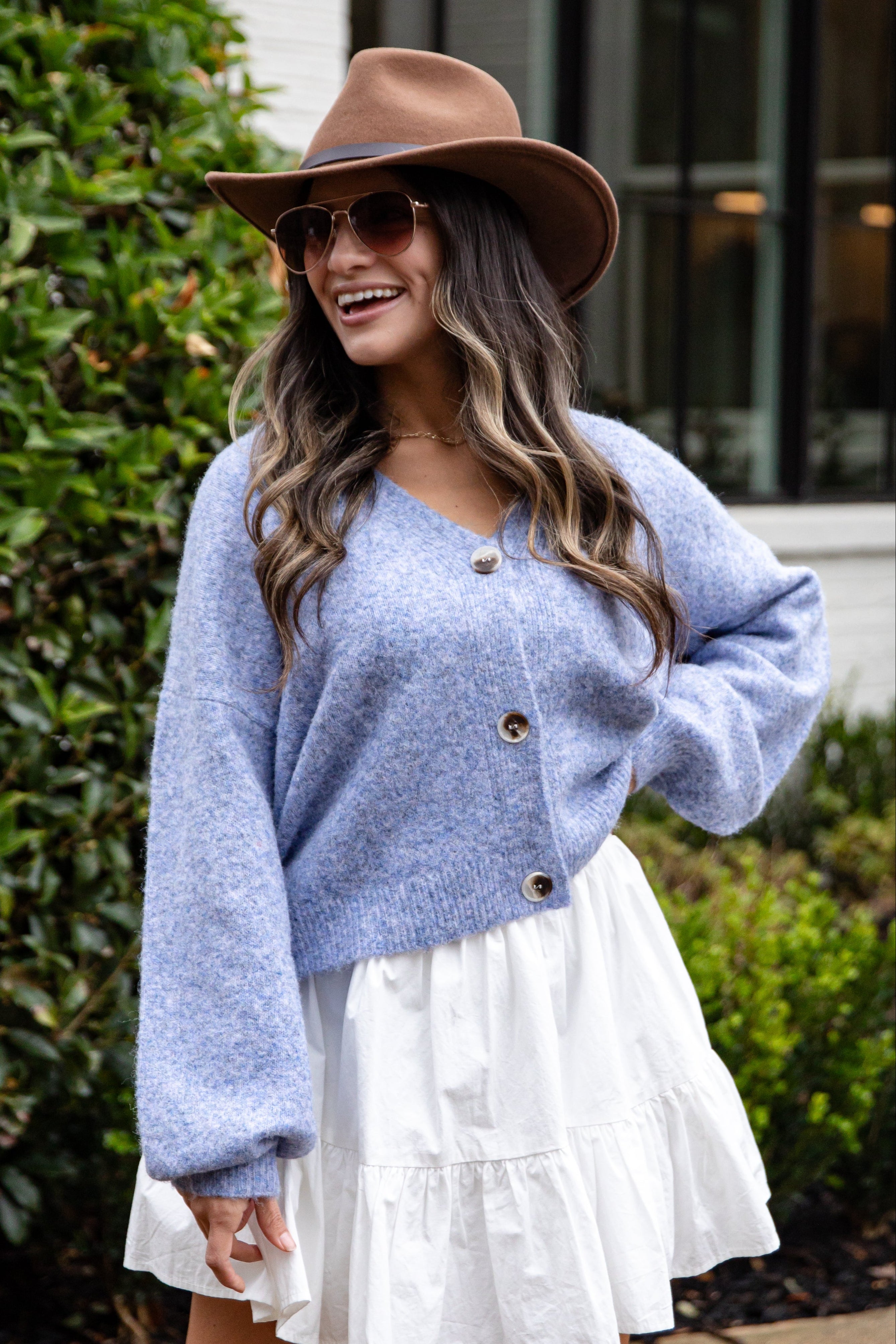 Woman wearing a blue cardigan and white dress with a brown hat outdoors.