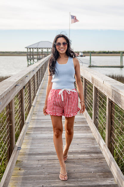 Woman walking on a wooden dock wearing a light blue sleeveless top and red checkered shorts.