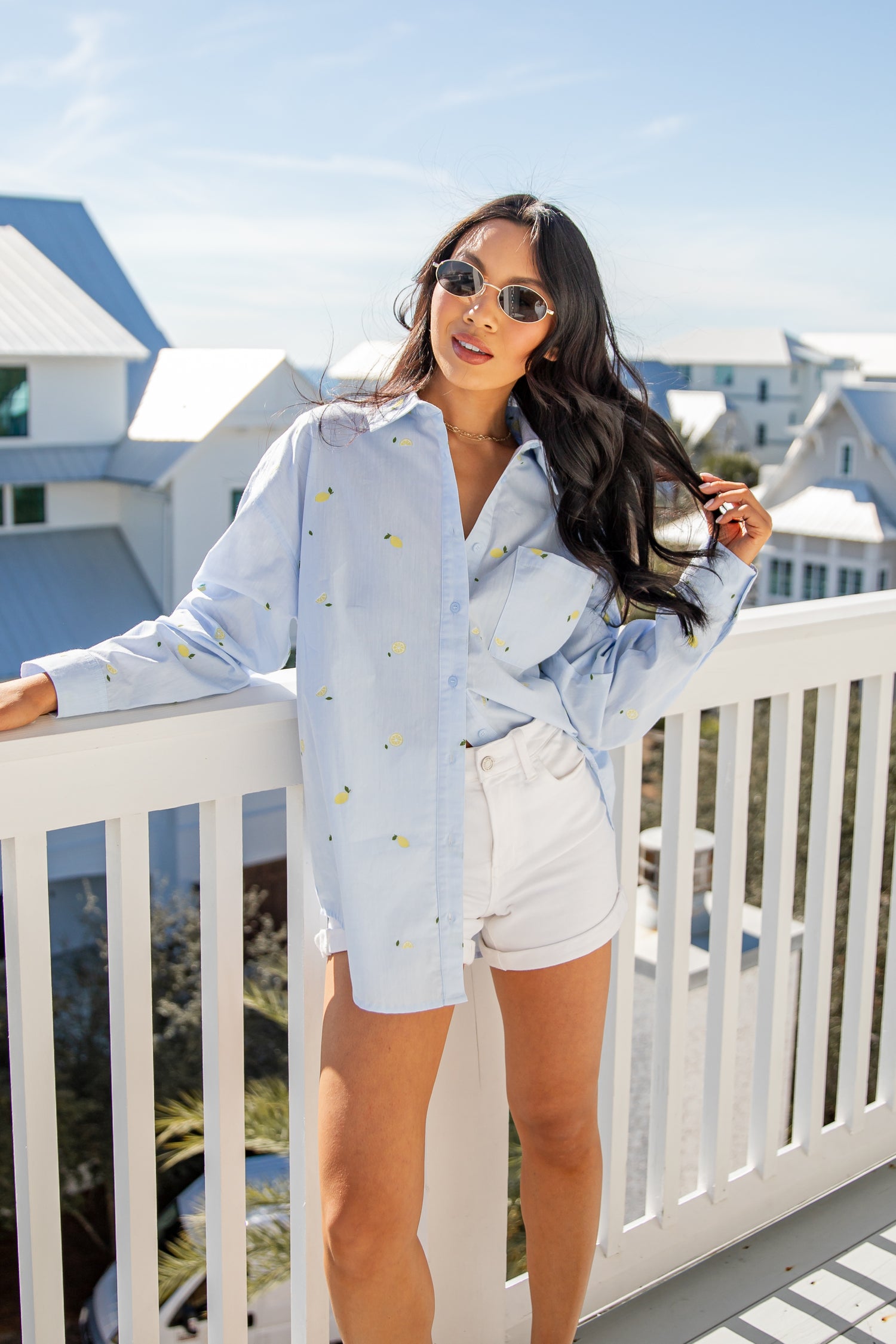 Woman in light blue shirt and white shorts standing on a balcony with a coastal background