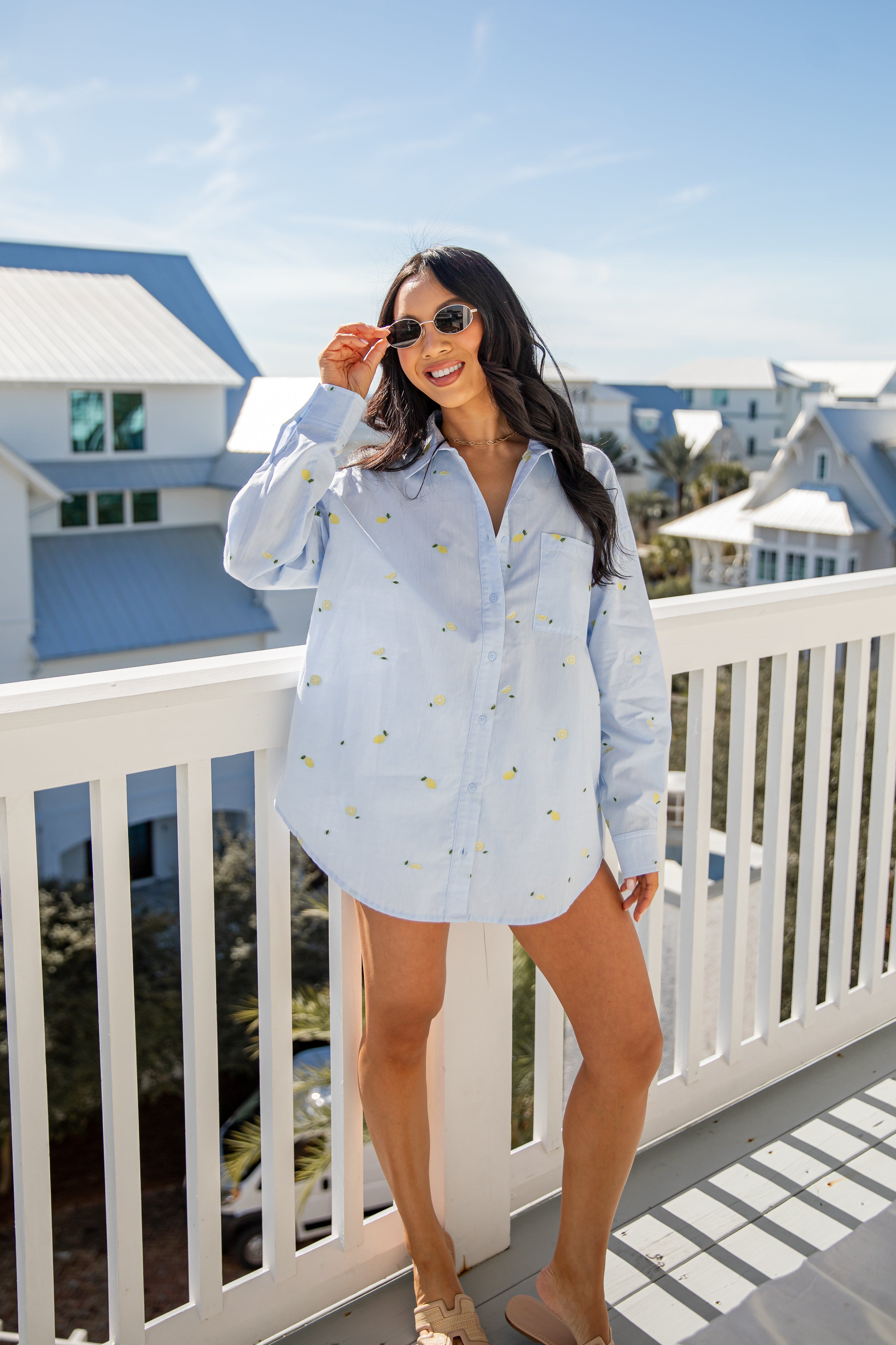 Woman in a light blue shirt with a pattern, standing on a balcony with a coastal view.