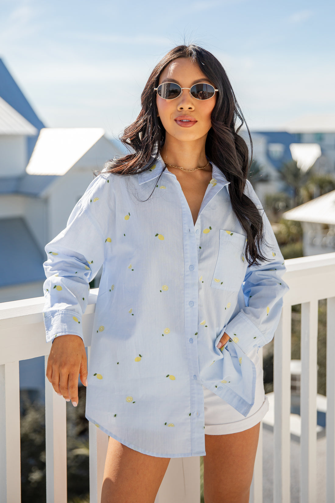 Woman wearing a light blue button-up shirt with yellow patterns and white shorts on a balcony.