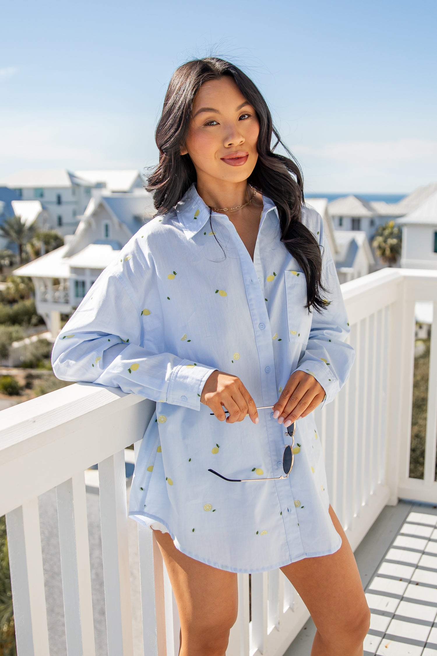 Woman wearing a light blue blouse with a pattern, standing on a balcony with a scenic background.