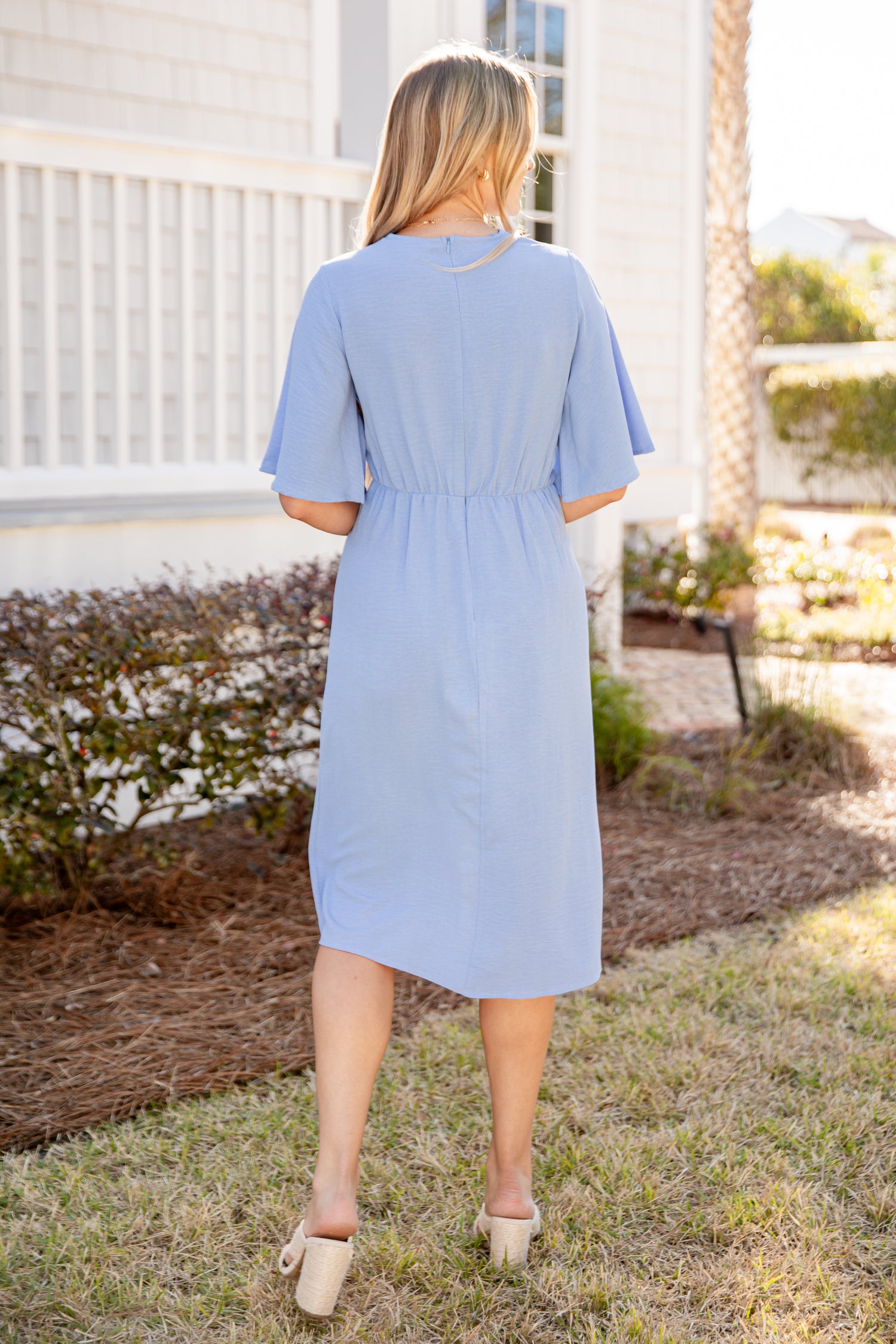 Woman wearing a light blue dress standing outdoors with a white building and palm tree in the background.