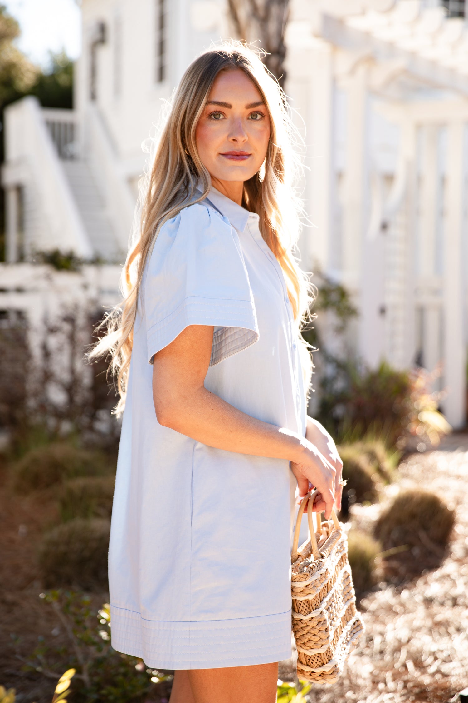 Woman in a light blue dress holding a straw bag outdoors.