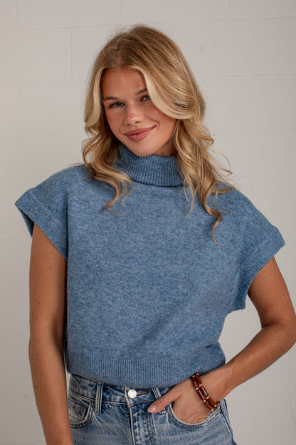 A woman wearing a heathered blue cropped knit turtleneck sweater with cap sleeves, paired with blue jeans, standing against a light gray brick wall.