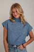 A woman wearing a heathered blue cropped knit turtleneck sweater with cap sleeves, paired with blue jeans, standing against a light gray brick wall.