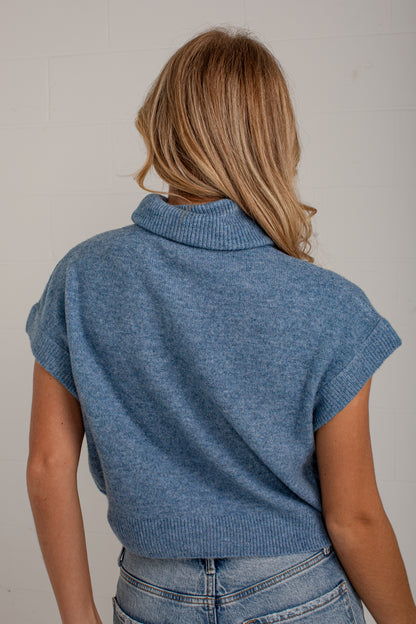A woman viewed from behind wearing a heathered blue short-sleeve mock neck knit sweater and light-wash jeans against a plain off-white wall.