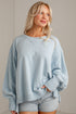 A woman wearing a light blue oversized crewneck sweatshirt and light-wash denim shorts, standing against a neutral off-white wall.