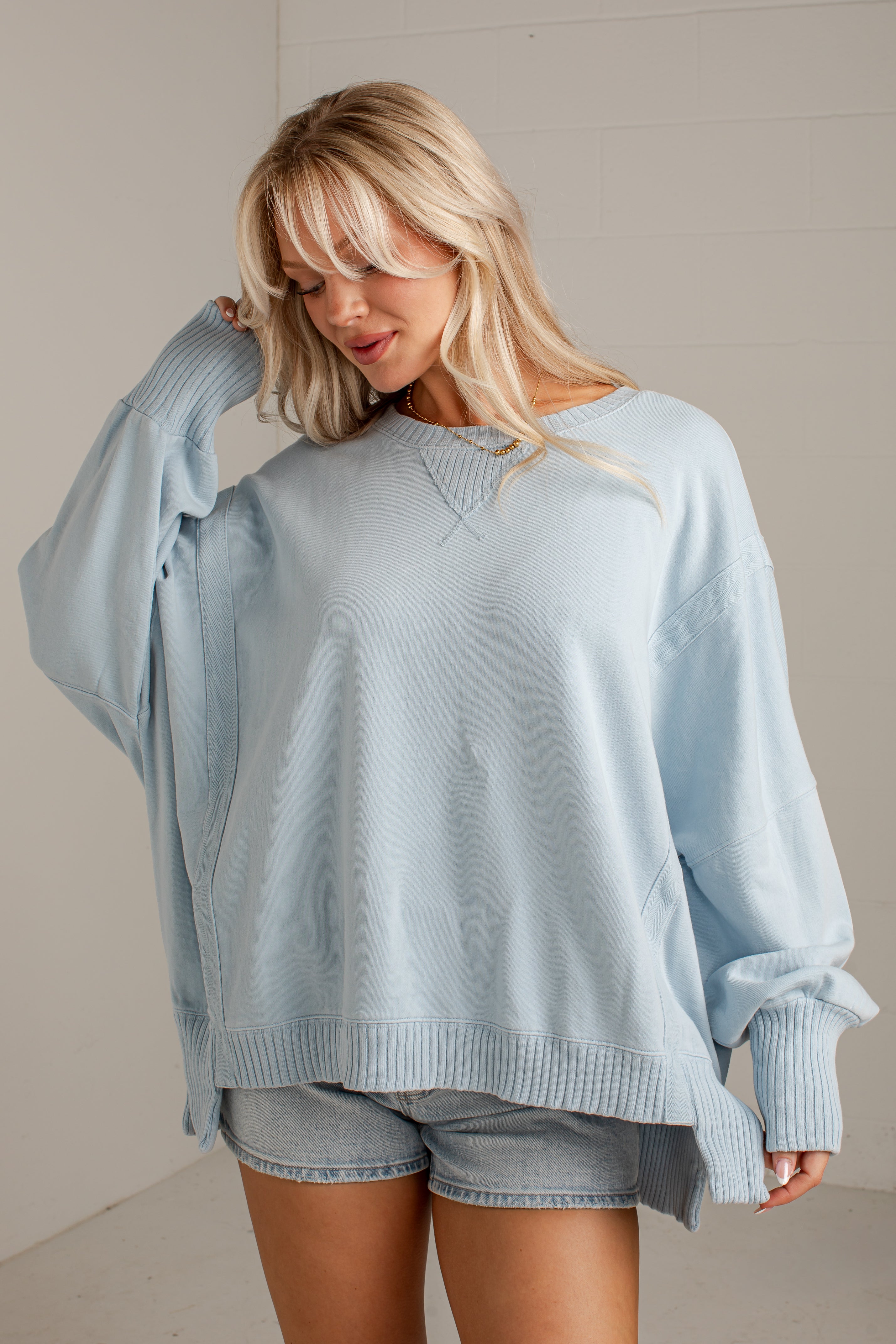A woman wearing an oversized light blue crewneck sweatshirt and denim shorts, posing in a bright indoor setting with a white paneled wall.
