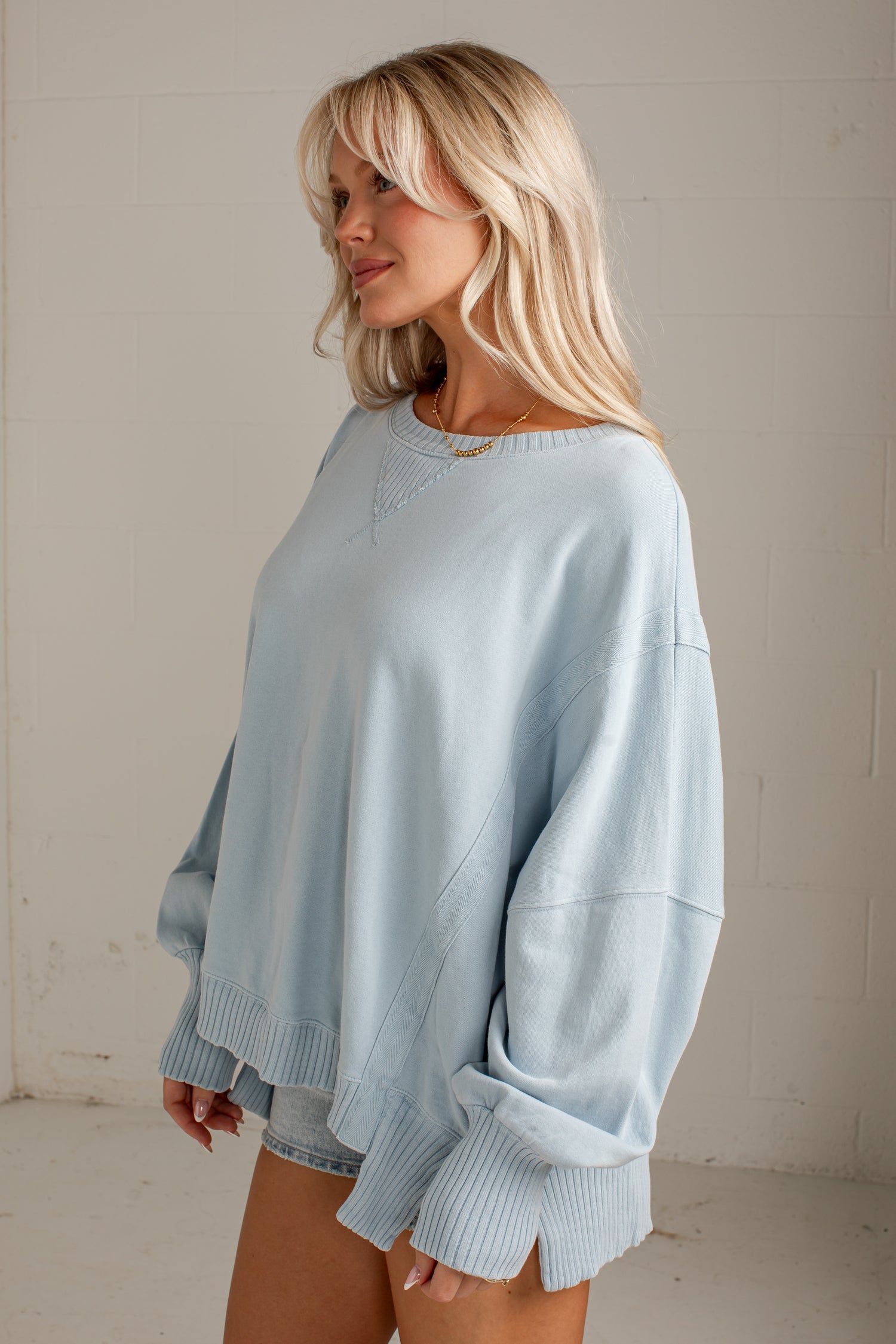 A woman wearing an oversized light blue sweatshirt with ribbed cuffs and hem, standing against a light-colored brick wall.