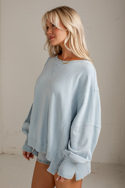 A woman wearing an oversized light blue sweatshirt with ribbed cuffs and hem, standing against a light-colored brick wall.