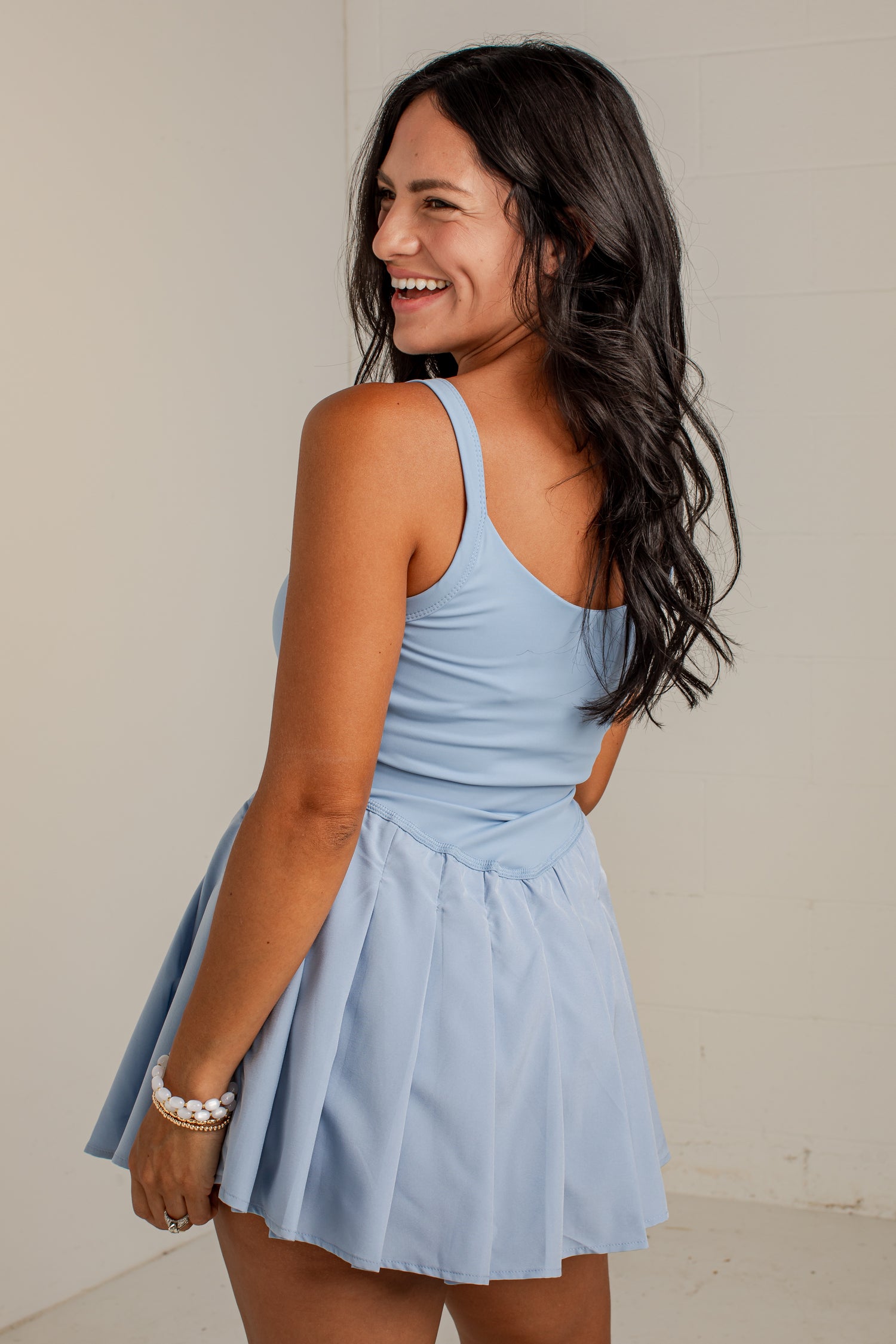 A woman with long dark hair is shown from the back wearing a light blue sleeveless mini dress with a pleated skirt, standing against a plain off-white wall.