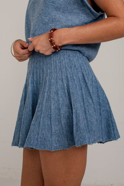 A person wearing a heathered blue knit sleeveless top and matching pleated mini skirt, shown from the chest down against a plain light wall.
