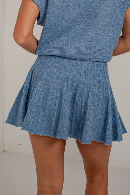 A person wearing a heathered blue ribbed knit sweater vest and matching pleated mini skirt, shown from the back against a light neutral wall.