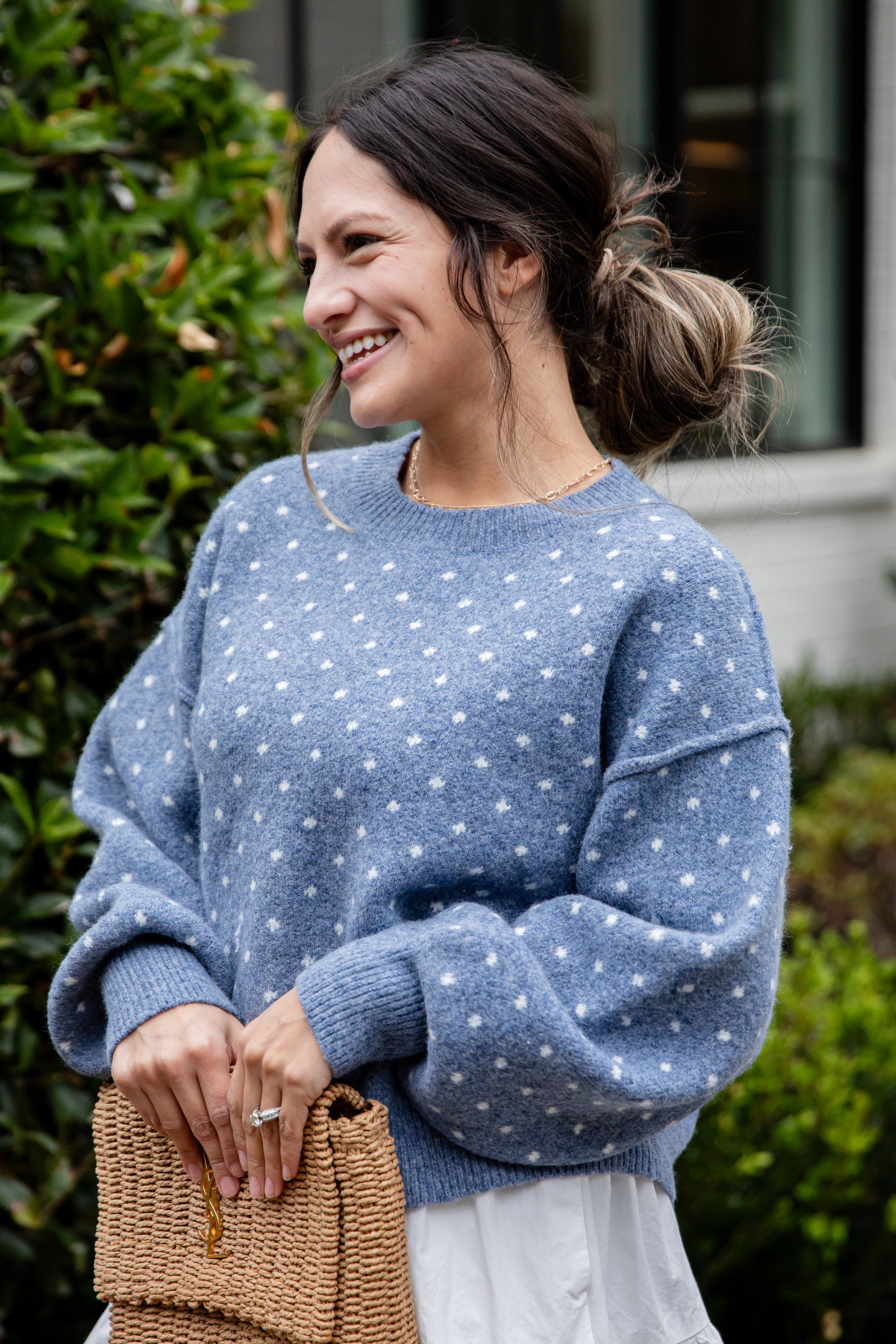 Woman wearing a blue polka dot sweater standing outdoors with greenery in the background