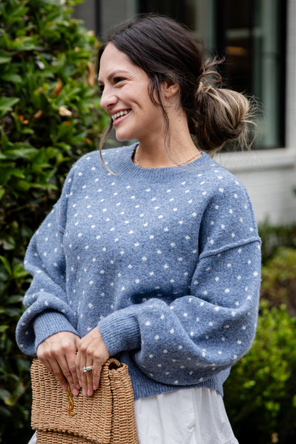Woman wearing a blue polka dot sweater standing outdoors with greenery in the background