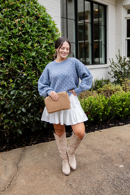Woman in a blue polka dot sweater, white skirt, and beige knee-high boots standing outdoors.