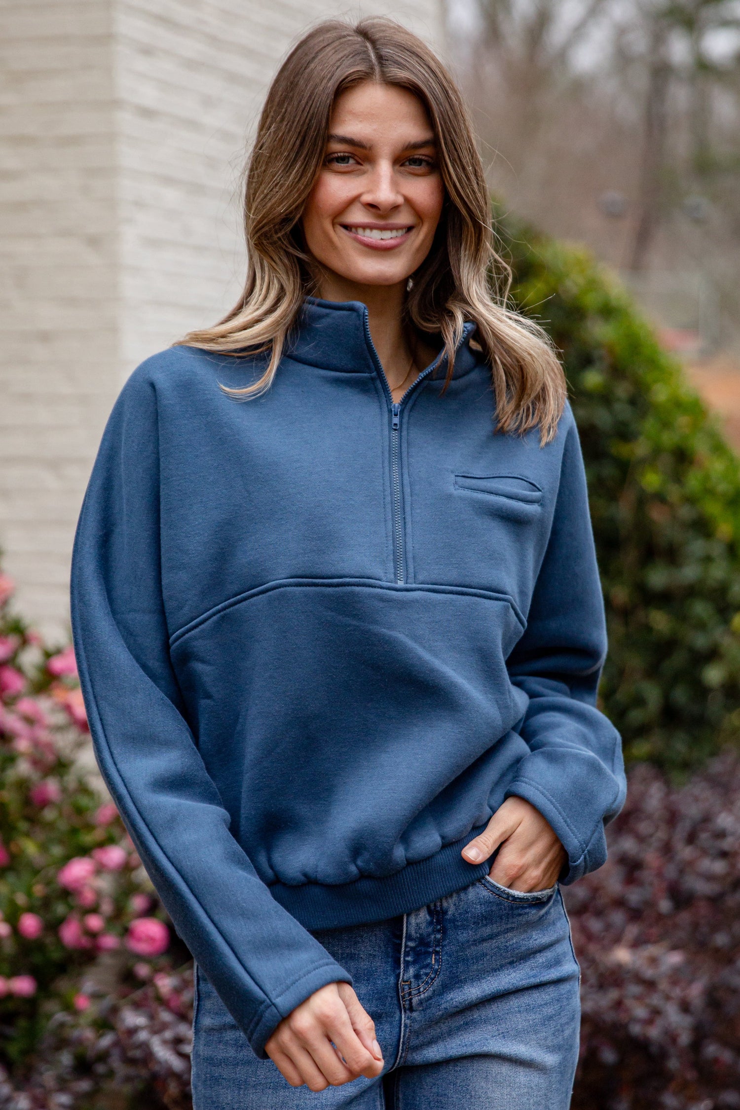 Woman wearing a blue pullover with a brick building and plants in the background