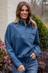 Woman wearing a blue pullover with a brick building and plants in the background