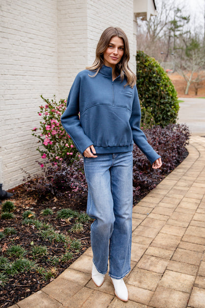 Woman wearing a blue sweatshirt and jeans standing on a sidewalk with a garden in the background