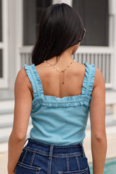 Woman wearing a light blue sleeveless top with ruffled straps and dark blue pants, standing outdoors.