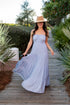 Woman in a long light blue dress and straw hat walking on a wooden path with greenery.