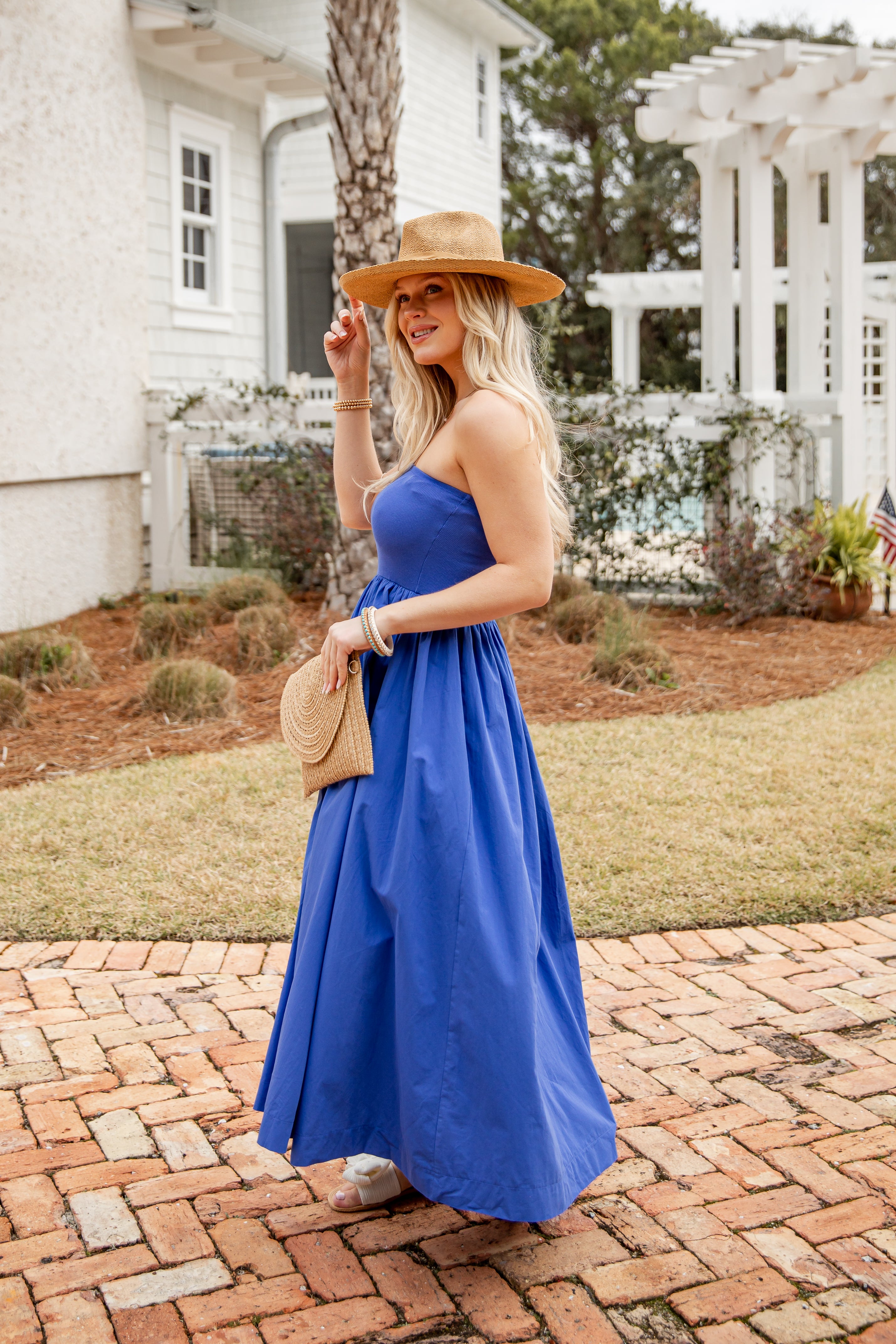Woman in a blue dress and hat standing on a brick path in front of a house.