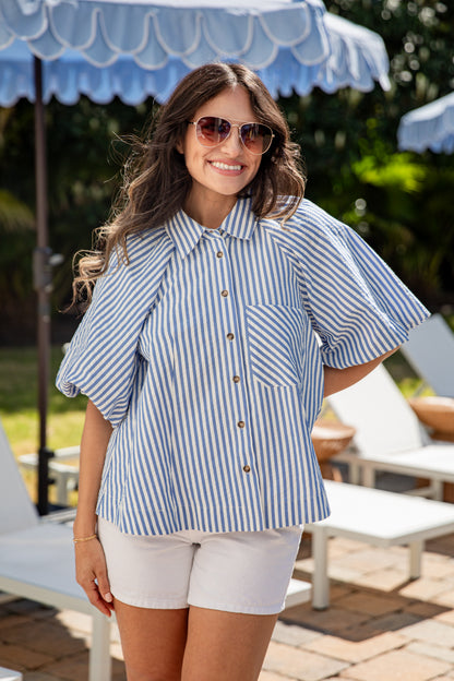 Woman wearing a blue and white striped shirt and sunglasses outdoors under an umbrella.