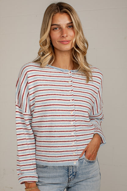 A woman wearing a fuzzy long-sleeve cardigan with thin red and white horizontal stripes, paired with light-wash blue jeans, standing against a light gray wall.