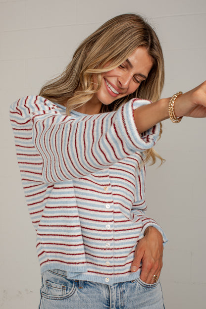 A woman wearing a fuzzy knit cardigan with horizontal red, white, and light blue stripes, paired with light-wash jeans, posing against a light gray wall.