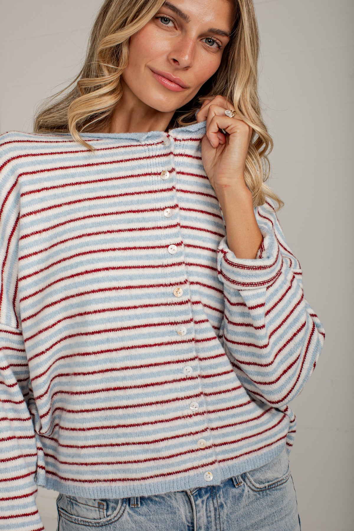 A woman wearing a fuzzy knit cardigan with horizontal red, white, and light blue stripes, paired with light-wash denim jeans, against a neutral gray background.