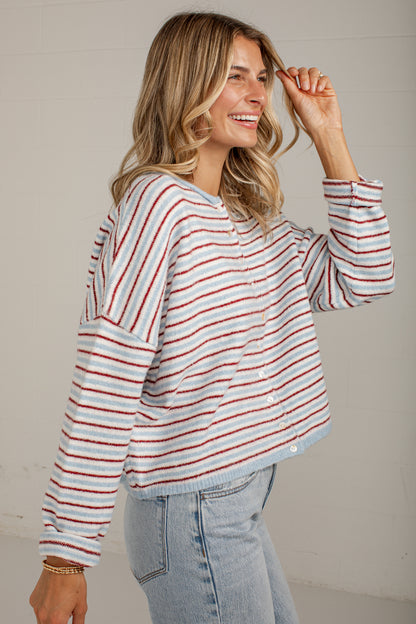 A woman wearing a long-sleeve striped knit top with red, white, and light blue horizontal stripes, paired with light-wash jeans, standing against a neutral light gray wall.